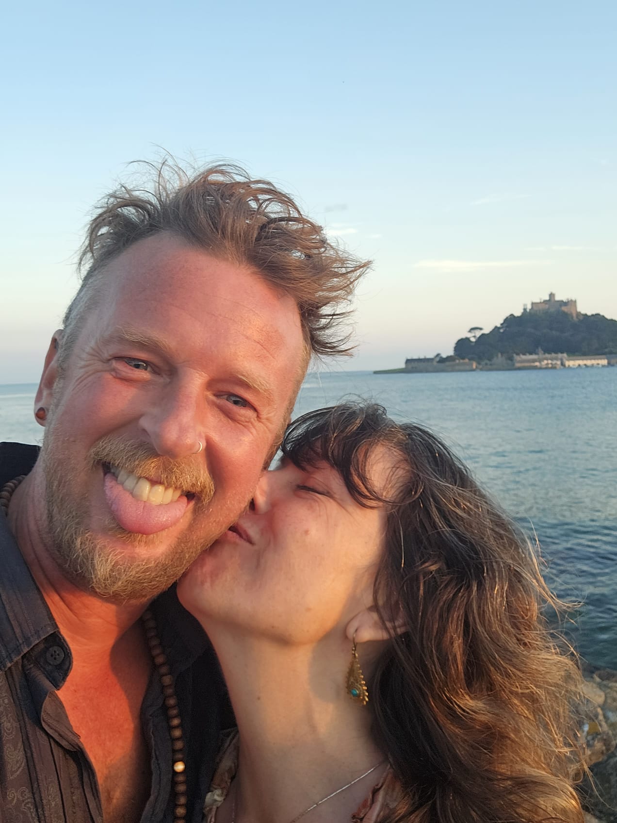 A man with tongue out and a woman — Cornwall wedding photography A man with tongue out and a woman kissing his cheek with a castle across the water for Cornwall wedding photography