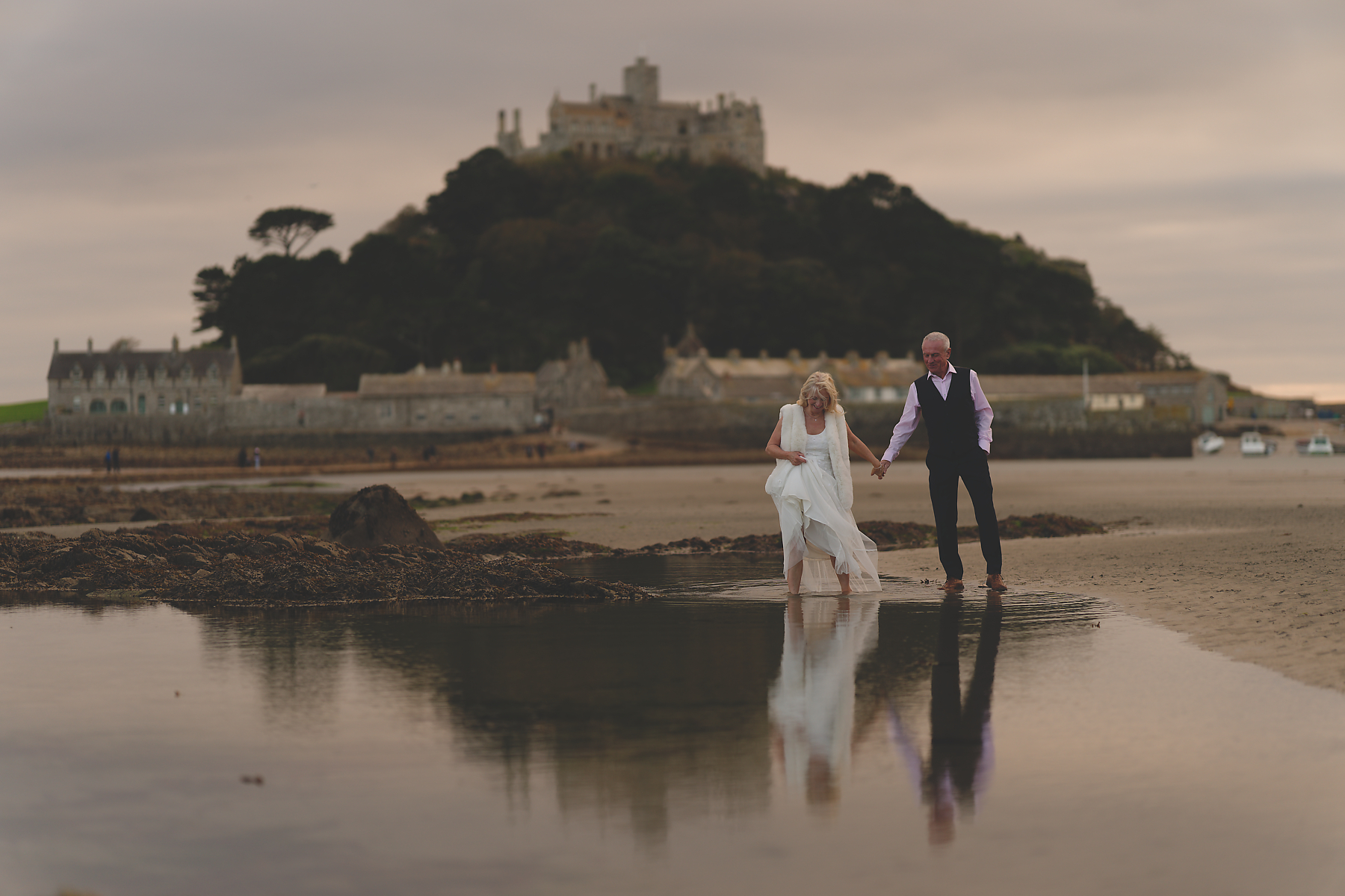 A bride and groom wade hand-in-hand through shallow — Cornwall wedding photography A bride and groom wade hand-in-hand through shallow water on a beach with a distant castle for Cornwall wedding photography